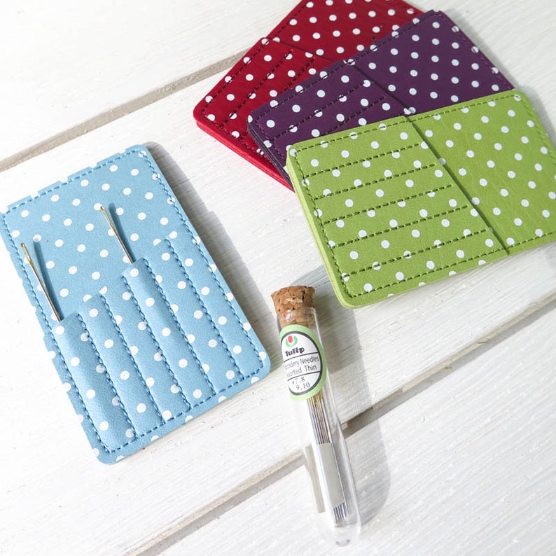 May include: A collection of sewing accessories, including a light blue needle case with white polka dots, a clear glass vial of needles, and several square coasters in red, purple, and green, all with white polka dots.
