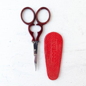 May include: Burgundy-handled embroidery scissors with a silver blade, alongside a red leather sheath. The scissors feature a decorative design and the blade is marked "PAKISTAN". The sheath is embossed with the word "KOOL RAY".