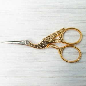 Pu&ograve; includere: Forbici da ricamo color oro con design a gru. Le forbici hanno una lama affilata e appuntita e un manico dettagliato a forma di uccello. I manici hanno due fori per le dita per un facile utilizzo. Le forbici sono su una superficie di legno bianco.