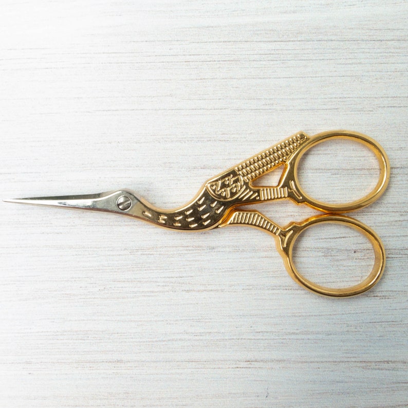 Pu&ograve; includere: Forbici da ricamo color oro con design a gru. Le forbici hanno una lama affilata e appuntita e un manico dettagliato a forma di uccello. I manici hanno due fori per le dita per un facile utilizzo. Le forbici sono su una superficie di legno bianco.
