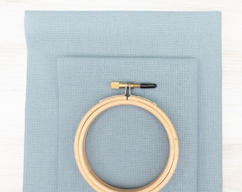 May include: A wooden embroidery hoop with a brass screw and black tip sits on light blue cross-stitch fabric. The fabric is neatly arranged, with the hoop positioned over the lower layer. The scene is captured from a top-down perspective.