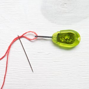 May include: A green Clover needle threader with a black needle and red thread. The threader has a clover design and a metal rod. The red thread is looped through the needle's eye, ready for sewing or embroidery.