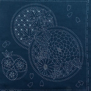 May include: Indigo blue textile with white Sashiko stitching. The design features three circular motifs with floral and geometric patterns, along with scattered petal shapes. The stitching creates a textured, handcrafted appearance, perfect for home decor or crafting projects.