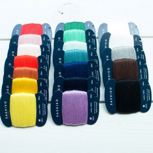 May include: A collection of Sashiko cotton thread spools in various colors, including red, yellow, green, blue, and purple. Each spool is labeled with text and measurements, and is made in Japan.