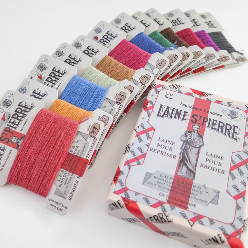 Puede incluir: Una colecci&oacute;n de carretes de hilo de bordar coloridos y una caja de hilo Laine St. Pierre. Los carretes est&aacute;n dispuestos en forma de abanico, mostrando varios tonos de rosa, azul, verde y marr&oacute;n. La caja presenta texto e ilustraciones en franc&eacute;s.