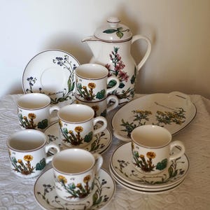 Puede incluir: Un juego de café de cerámica que incluye una cafetera alta con tapa, cinco tazas, cinco platillos y una bandeja ovalada. Cada pieza está decorada con ilustraciones botánicas de flores amarillas y azules, incluyendo el texto Myosotis palustris.