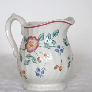 Peut inclure: Un pot à lait en céramique blanche orné d'un motif floral délicat avec des fleurs roses, des feuilles vertes, de petites fleurs bleues et des baies orange. Le pot présente un corps cannelé, une anse incurvée et un fin liseré rouge sur le bord.