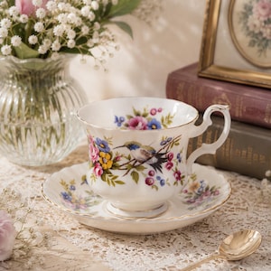 Op de afbeelding: Een witte porseleinen theekop en schotel met een kleurrijk bloemmotief van roze rozen, blauwe bloemen en een centrale illustratie van een kolibrie. De theekop heeft een geschulpte rand met een gouden bies en een decoratief handvat.