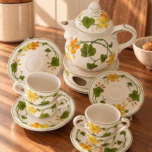 Op de afbeelding: Een wit keramieken theeservies met een handgeschilderd patroon van groene ranken met gele en oranje bloemen. De set bevat een theepot met deksel op een theelichthouder, twee theekopjes met schotels en twee extra schotels met bloemmotieven.