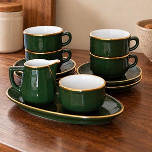 Può includere: Un set di stoviglie in ceramica verde scuro con interni bianchi e bordi dorati. La collezione comprende quattro tazze impilate su piattini, una piccola lattiera e una ciotola, il tutto disposto su un vassoio da portata di forma ovale.