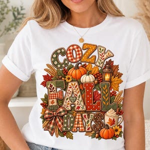 May include: A white short-sleeved t-shirt featuring a colourful graphic print with the text COZY FALL DAYS. The design includes pumpkins, autumn leaves, a lantern, a plaid bow, and a mug of hot beverage, all arranged in a decorative seasonal pattern.