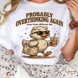 May include: A white crewneck t-shirt featuring a graphic of a duck wearing sunglasses, carrying a tote bag, and holding a coffee cup. The text reads PROBABLY OVERTHINKING AGAIN and same brain different day. The shirt is paired with leopard print pants.