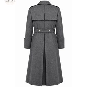 May include: A charcoal gray long coat featuring a back storm flap, a belted waist with two silver-toned buttons, and wide cuffs. The coat has a structured collar and a long, pleated back vent. The AVELUNE CLOTHING logo is visible in the top corner.