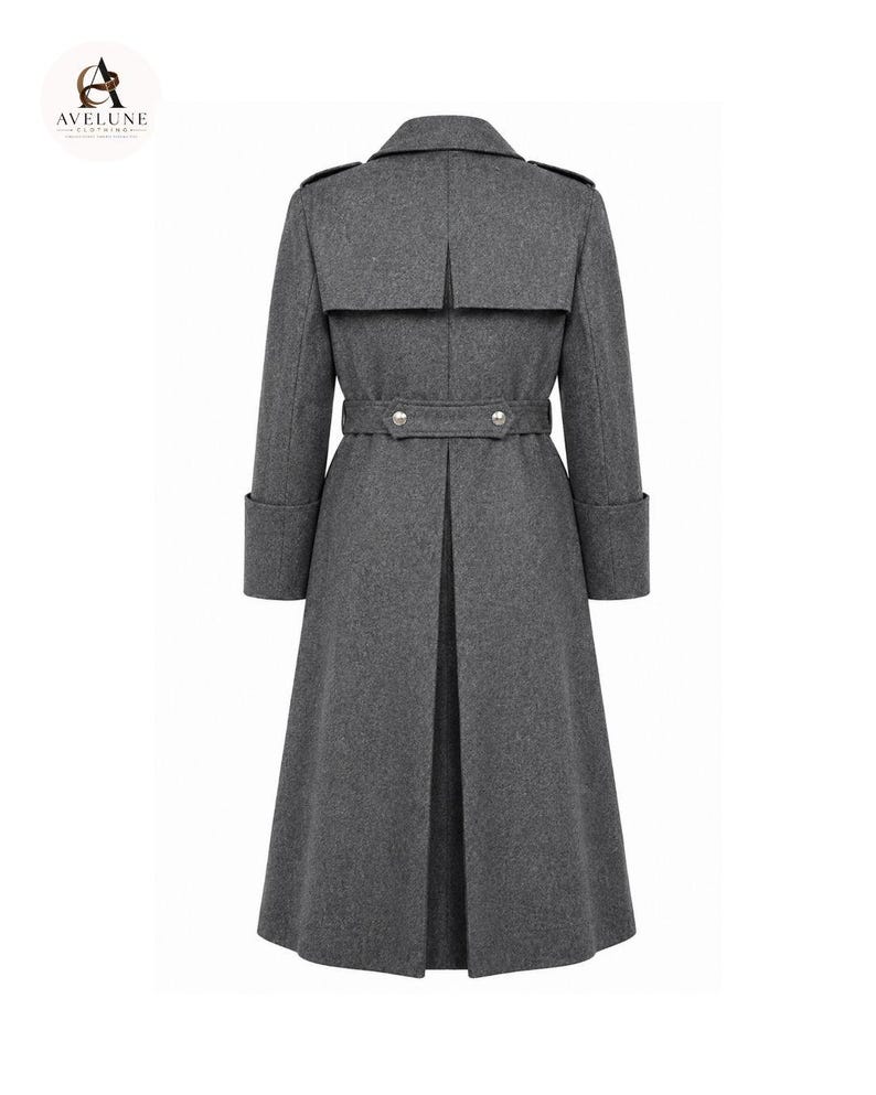 May include: A charcoal gray long coat featuring a back storm flap, a belted waist with two silver-toned buttons, and wide cuffs. The coat has a structured collar and a long, pleated back vent. The AVELUNE CLOTHING logo is visible in the top corner.