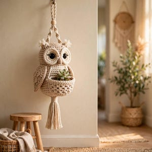 Puede incluir: Un colgante de pared de búho tejido en crochet de color beige con un compartimento central en forma de cesta que contiene una pequeña planta suculenta verde. El búho tiene ojos negros grandes, mechones en las orejas y una borla larga.