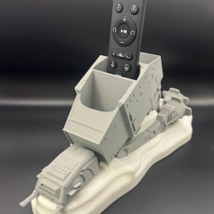 May include: A gray 3D-printed walker-shaped remote control holder featuring a hollow rectangular compartment for storage. The holder is mounted on a white, uneven, snow-like base. A black rectangular remote control with various buttons is inserted inside.
