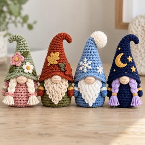 May include: Four handmade crochet gnomes standing in a row. The first is green with pink flowers and blonde braids. The second is orange with autumn leaves and a beard. The third is blue with white snowflakes and a beard. The fourth is dark blue with a moon and stars and purple braids.