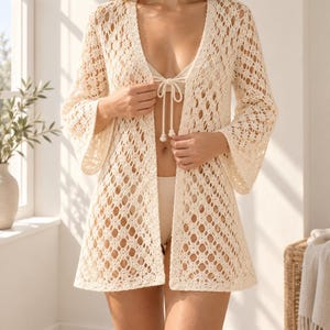 May include: An off-white open-front cardigan featuring an intricate crochet pattern with a geometric diamond design, long flared sleeves, and a straight hemline. The garment is worn over a matching off-white bikini top and bottom set.