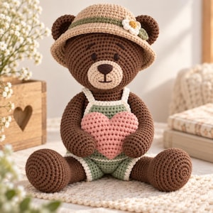Puede incluir: Un oso de peluche tejido a ganchillo de color marrón oscuro que lleva un sombrero de sol beige con una pequeña flor blanca y un pelele verde claro con ribete blanco. El oso sostiene un corazón tejido rosa suave contra su pecho.
