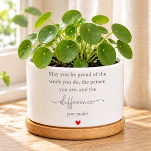 May include: A white ceramic cylindrical planter with a bamboo saucer base containing a green Pilea peperomioides plant. The planter features black printed text that reads: May you be proud of the work you do, the person you are, and the difference you make, with a small red heart icon below the text.