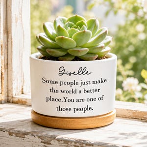 May include: A white ceramic planter containing a green succulent plant. The planter features the black printed text: Giselle, Some people just make the world a better place. You are one of those people. It sits on a circular light wood base.