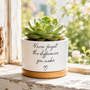 May include: A white ceramic cylindrical planter with a light wood saucer base, containing a green succulent plant. The front of the planter features the black printed text "Never forget the difference you make" above a small hand-drawn black heart.