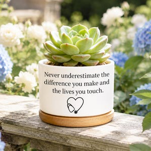 Könnte beinhalten: Ein zylindrischer Übertopf aus weißer Keramik mit hellem Holzsockel, der eine grüne Sukkulente enthält. Der Topf ist mit dem schwarzen Text Never underestimate the difference you make and the lives you touch sowie einem Herz- und Stethoskop-Symbol bedruckt.