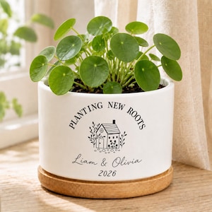 May include: A white cylindrical ceramic planter with a wooden saucer base containing a green Pilea peperomioides plant. The planter features black text reading "PLANTING NEW ROOTS", "Liam & Olivia", "2026", and a graphic illustration of a small house.