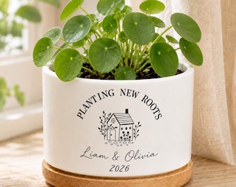 Könnte beinhalten: Ein zylindrischer weißer Keramik-Pflanztopf mit einem hölzernen Untersetzer, der eine grüne Pilea peperomioides enthält. Der Topf ist mit dem schwarzen Text "PLANTING NEW ROOTS", "Liam & Olivia", "2026" und einer Grafik eines kleinen Hauses bedruckt.