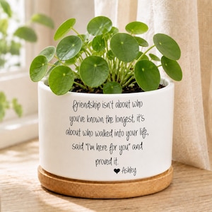 May include: A white ceramic cylindrical planter with a light wood base containing a Pilea peperomioides plant. The planter features black printed text: "friendship isn't about who you've known the longest, it's about who walked into your life, said "I'm here for you" and proved it. ♥ Ashley".