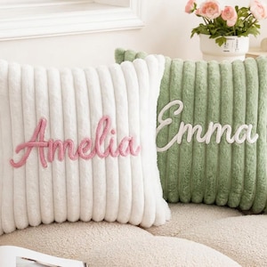 Puede incluir: Dos cojines cuadrados con textura acanalada y afelpada. El cojín blanco presenta el nombre Amelia bordado en letra cursiva rosa, mientras que el cojín verde salvia muestra el nombre Emma bordado en letra cursiva blanca en el centro.