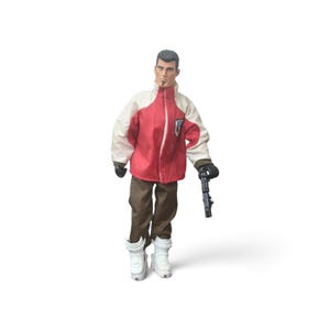 May include: A posable action figure with short dark hair and a goatee, wearing a red and cream-colored jacket with a small emblem, brown trousers, and white high-top boots. The figure holds a black toy pistol in one hand and wears black gloves.