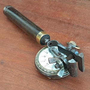 Può includere: Un antico strumento da orologiaio con manico cilindrico scuro e ghiera in ottone, collegato a un meccanismo di serraggio in metallo. Il morsetto è posizionato sopra un quadrante circolare bianco con numeri neri, fissato da un dado a farfalla.