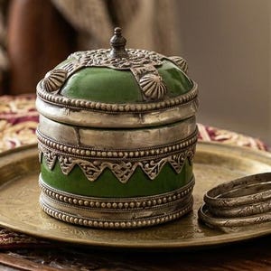 Peut inclure: Un récipient rond vert et argenté avec couvercle est posé sur un plateau en laiton avec deux bracelets en argent. À côté, une petite bouteille en verre transparent remplie de perles claires, un carnet en cuir marron et un crayon.