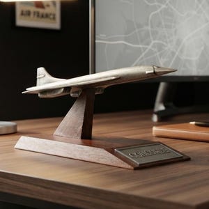 Pode incluir: Um modelo de metal prateado de um avião Concorde montado num suporte de madeira castanho-escuro com uma placa metálica retangular. O modelo está posicionado sobre uma secretária de madeira ao lado de um candeeiro, um caderno, uma caneta e uma caneca preta.