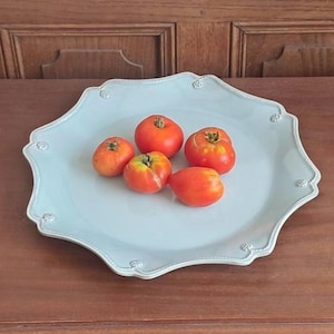 May include: A light blue ceramic serving platter with a scalloped edge and small embossed circular details along the rim. Five fresh, round red tomatoes with green stems are arranged in the centre of the smooth, glossy surface of the plate.