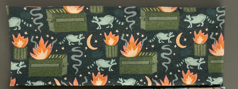 Puede incluir: Una tela verde oscura con un patr&oacute;n repetido de mapaches, fuego y botes de basura. Los mapaches son negros y blancos, el fuego es naranja y amarillo, y los botes de basura son verdes. El fondo es verde oscuro.