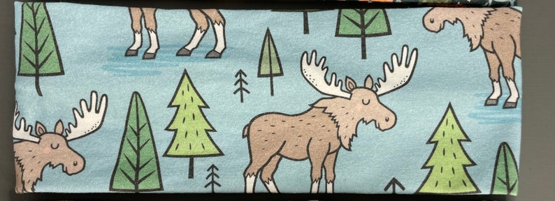 Puede incluir: Una diadema de tela azul con un patr&oacute;n repetido de alces, &aacute;rboles y ciervos. Los alces son marrones con grandes cuernos, los &aacute;rboles son verdes y los ciervos son marrones con manchas blancas.