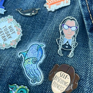 Op de afbeelding: Een verzameling emaille pins met verschillende ontwerpen, waaronder een narwal, een inktvis, een bank, een vrouw met een bril, een prinses en een krans met de tekst "Not Today Satan". De pins zijn bevestigd aan een blauw denim jack.