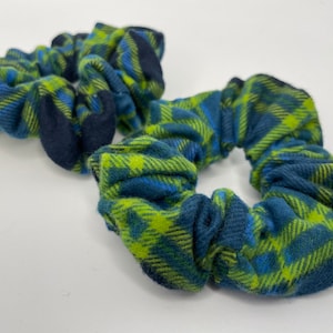 May include: Two scrunchies with a blue, green, and navy plaid pattern. The fabric is soft and gathered, creating a textured appearance. The scrunchies are designed for hair accessories.