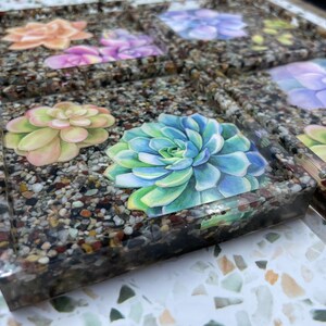 Puede incluir: Un juego de posavasos cuadrados de resina con textura de guijarros. Cada posavasos presenta una ilustración de acuarela de una planta suculenta en tonos verdes, azules, rosas y morados. Los posavasos están dispuestos en forma de cuadrícula.