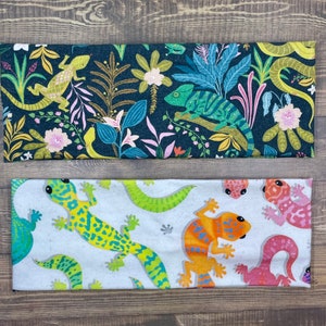 May include: Two headbands with reptile designs. One has a dark teal background with lizards, chameleons, and floral patterns. The other features a light grey background with colourful geckos in green, orange, and pink.