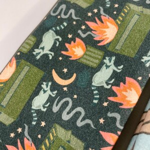 Puede incluir: Una tela de color verde oscuro, azul y naranja con un patr&oacute;n repetido de mapaches, llamas y estrellas. La tela tiene una superficie texturizada.