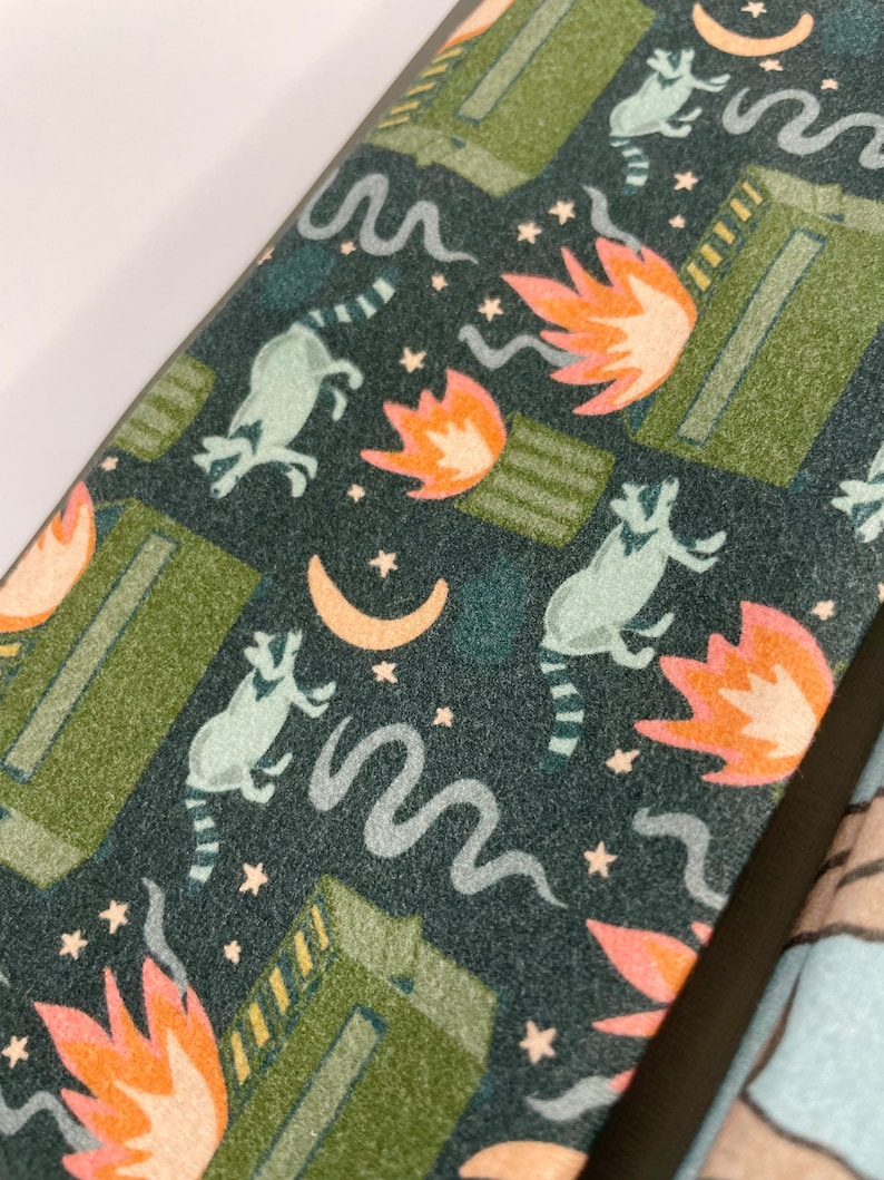 Puede incluir: Una tela de color verde oscuro, azul y naranja con un patr&oacute;n repetido de mapaches, llamas y estrellas. La tela tiene una superficie texturizada.