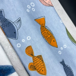 以下が含まれることがあります： オレンジとブルーの魚、白い泡、ピンクとブラウンの円形のパターンが繰り返されるライトブルーの生地。