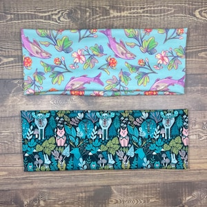 May include: Two fabric headbands with different patterns. The top headband is light blue with a white and green floral pattern and a salmon fish design. The bottom headband is black with a green and white woodland creature pattern.