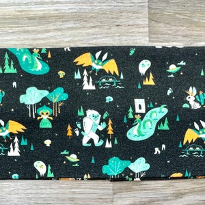 Könnte beinhalten: Schwarzer Stoff mit einem sich wiederholenden Muster aus grünen, weißen und gelben Waldtieren, darunter ein Yeti, ein Kaninchen, ein Drache und ein Flugwesen. Der Hintergrund ist schwarz mit einer dezenten Textur.