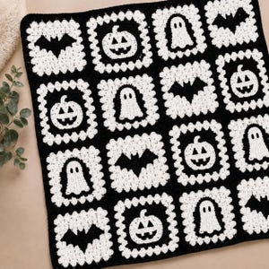 May include: A square crochet blanket featuring a grid of sixteen alternating black and white squares. Each square displays a motif of a ghost, a bat, or a jack-o'-lantern. The blanket is constructed with a textured yarn pattern throughout the design.