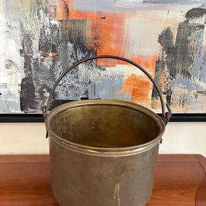 May include: A cylindrical brass bucket with a smooth, weathered patina and a dark metal swing handle attached to two side loops. The vessel features a wide, open top and a rounded base, showing signs of oxidation and surface discoloration.