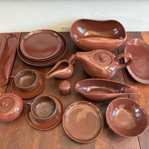 May include: A collection of brown ceramic dinnerware including a teapot, creamer, sugar bowl, two cups with saucers, two plates, a large serving bowl, a gravy boat, a small shaker, and two elongated serving platters with a smooth finish.